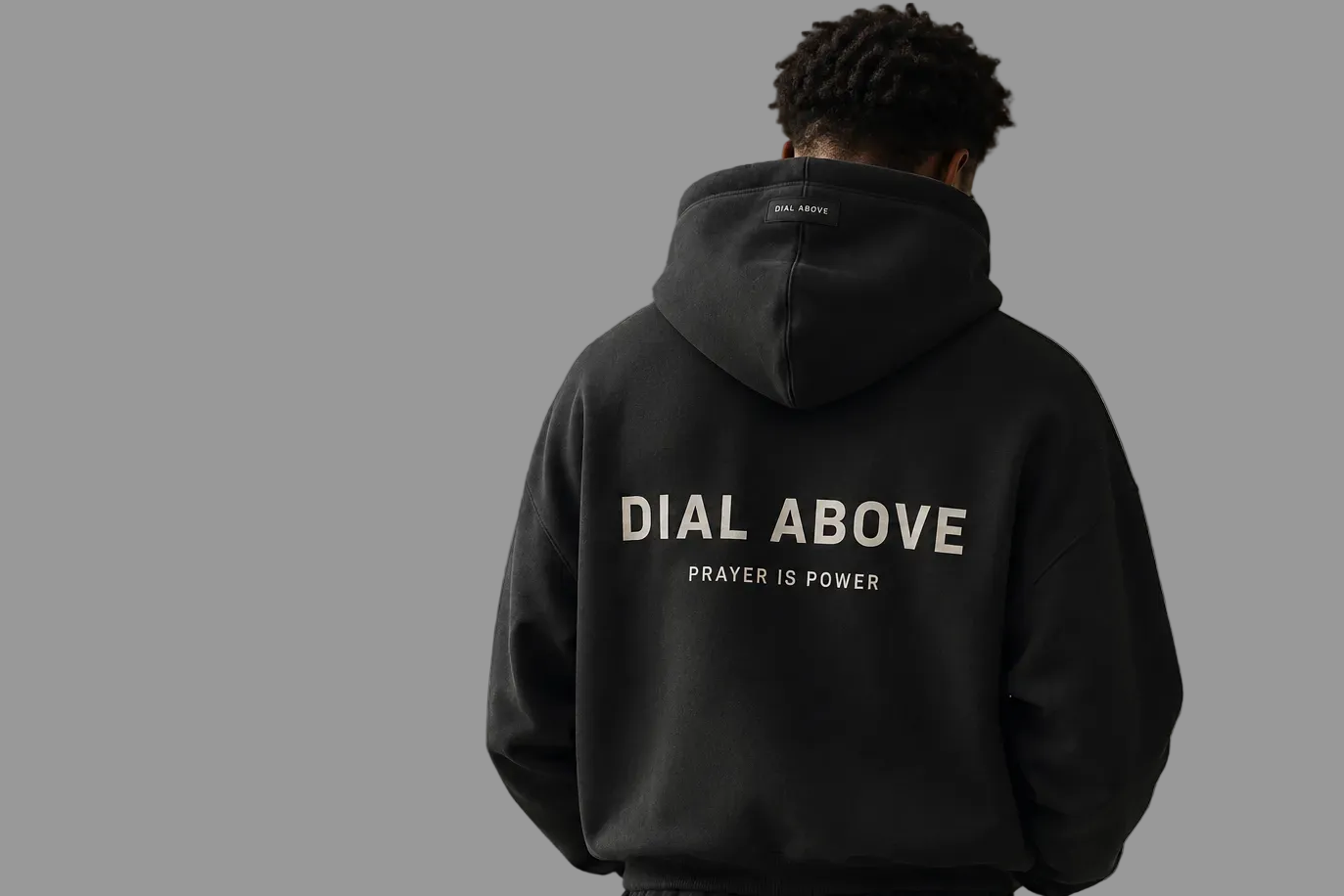Person wearing a black hoodie with text Prayer is Power Dial Above on the back against a beige background